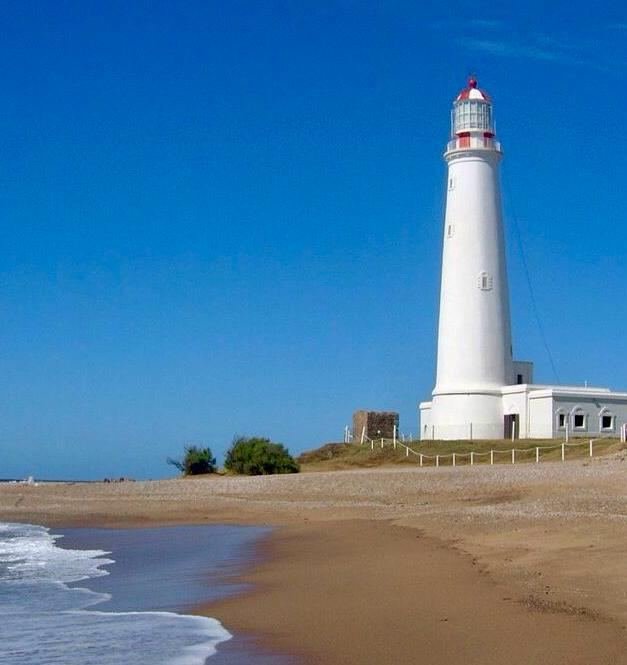 Faro blanco con cúpula roja sobre la costa, con olas en primer plano